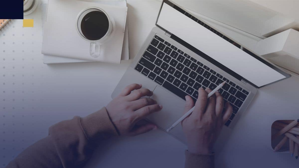 Manos escribiendo en laptop con café y cuadernos para redactar monografía académica.