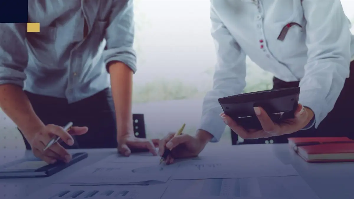 Dos personas planificando un proyecto académico, revisando documentos y utilizando una calculadora sobre una mesa de trabajo.