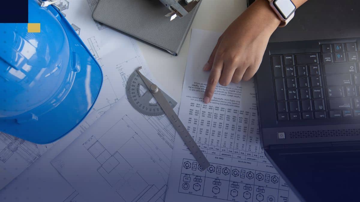 Planos de Ingeniería Mecánica sobre mesa con casco de seguridad, calibrador y laptop para desarrollo de tesis de grado.