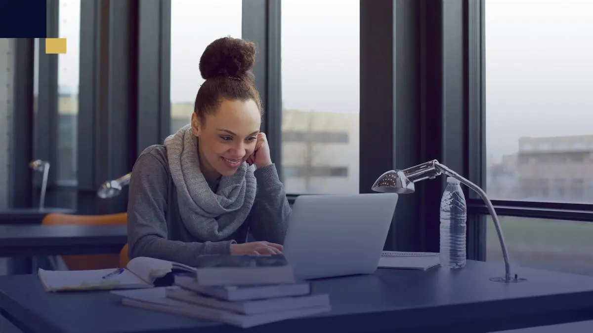 Estudiante universitaria trabajando en una biblioteca, usando laptop y rodeada de libros y apuntes para organizar fichas bibliográficas.