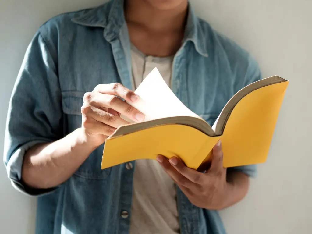 Revisión de literatura para el marco metodológico: estudiante consultando fuentes académicas Estudiante universitario hojeando un libro amarillo, revisando información relevante para la elaboración del marco metodológico de su tesis.