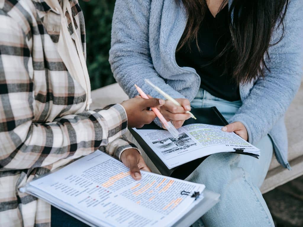 Estudiantes revisando y subrayando documentos para comparar tipos de tesis y enfoques académicos Dos estudiantes universitarias sentadas al aire libre revisando apuntes y subrayando información en documentos impresos, colaborando en la organización y análisis de diferentes tipos de tesis.