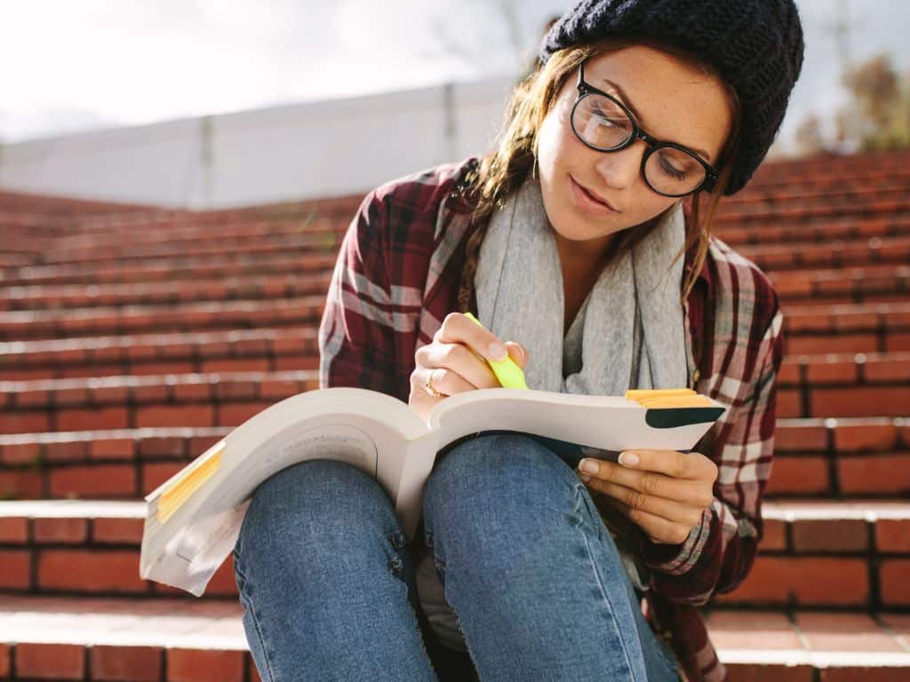 Estudiante universitaria subrayando y tomando notas en libro durante sesión de estudio al aire libre Estudiante universitaria sentada en gradas al aire libre, subrayando y tomando notas en un libro abierto durante una sesión de estudio.
