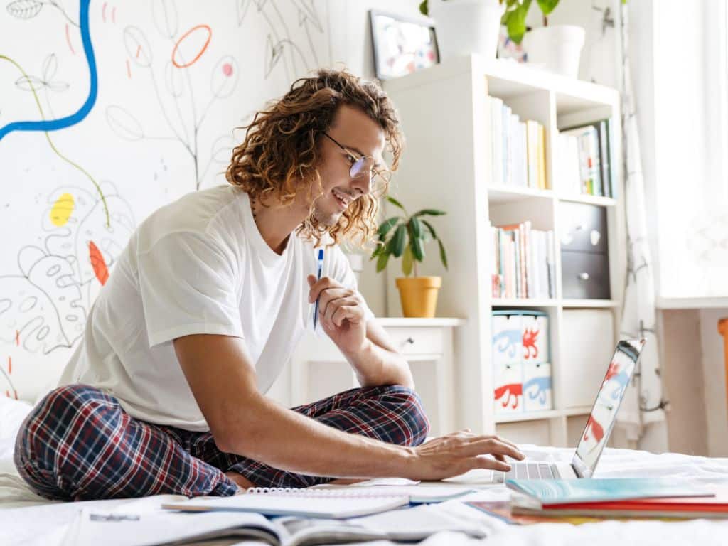 Cómo hacer un resumen académico: estudiante redactando con laptop y libros en su habitación Estudiante universitario redactando un resumen académico en su habitación, usando laptop y rodeado de libros y apuntes.
