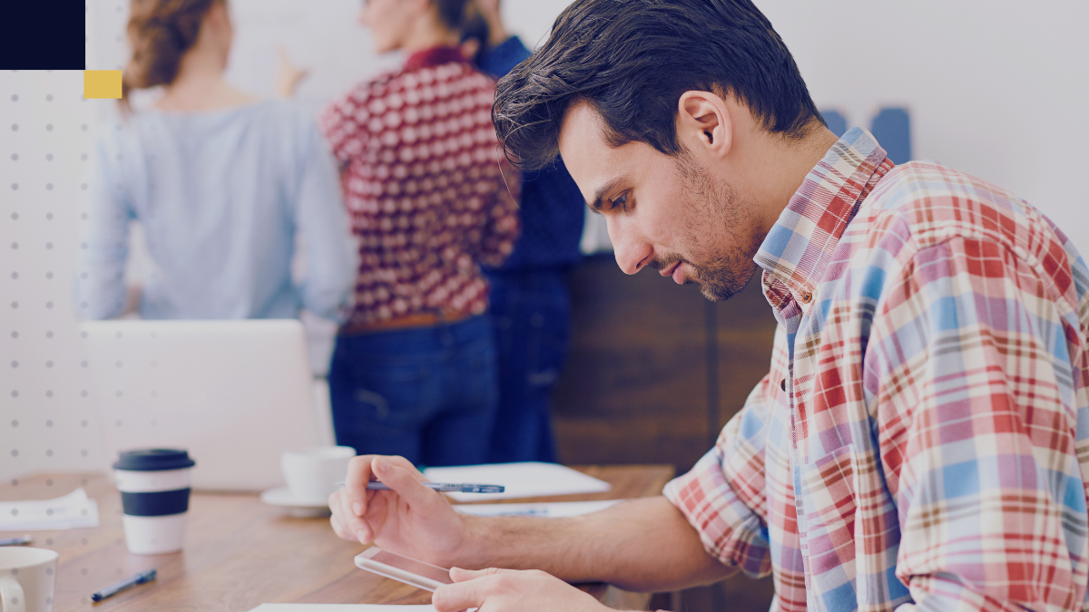 Joven profesional revisando documentos y tomando notas en una tablet en un entorno de oficina, mientras al fondo un grupo de colegas discute estrategias, representando el análisis colaborativo en estudios de caso empresariales.
