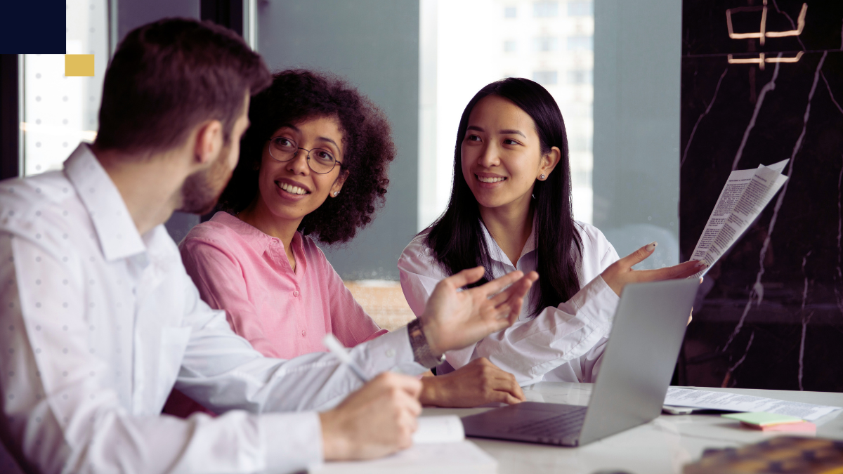 Tres personas adultas dialogando y revisando documentos frente a una laptop, colaborando en la planificación de un proyecto académico multidisciplinario.